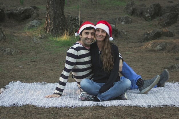 Porträt eines glücklichen Paares mit roten Weihnachtsmützen, das auf einer Decke im Pinienwaldpark sitzt?
