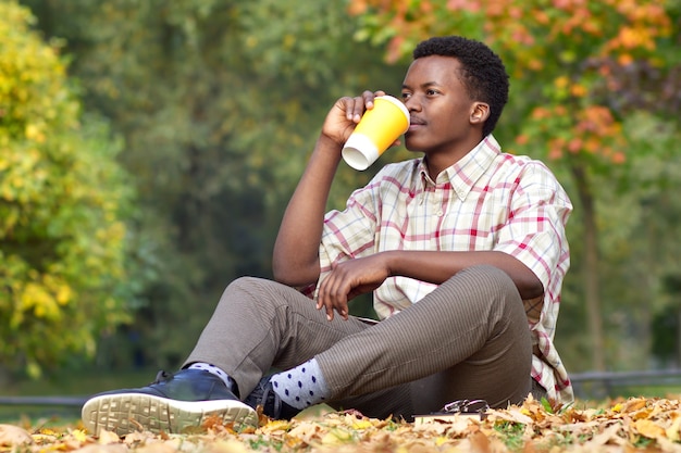 Porträt eines glücklichen schwarzen afrikanischen afroamerikanischen jungen ethnischen gutaussehenden Mannes, der auf dem Gras sitzt