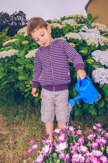 Porträt eines glücklichen süßen Jungen, der Blumen im Garten mit einer blauen Gießkanne gießt