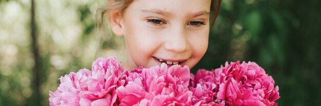 Porträt eines glücklichen süßen kleinen kaukasischen siebenjährigen Mädchens, das in Händen hält und riecht und einen Strauß rosa Pfingstrosenblumen in voller Blüte auf dem Hintergrund der Natur genießt. Banner