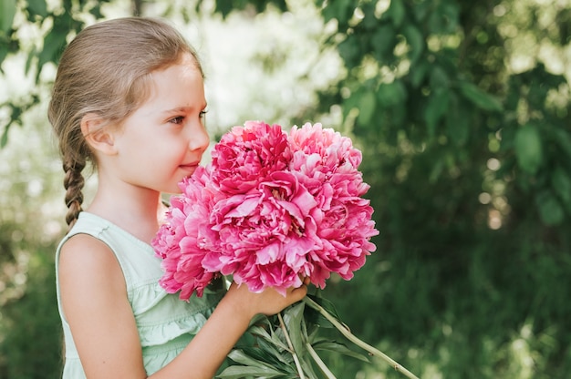 Porträt eines glücklichen süßen kleinen kaukasischen siebenjährigen Mädchens, das in Händen hält und riecht und einen Strauß rosa Pfingstrosenblumen in voller Blüte auf dem Hintergrund der Natur genießt