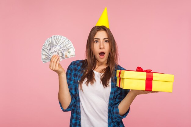 Porträt eines glücklichen, überraschten Mädchens mit lustigem Partykegel auf dem Kopf, das eine Geschenkbox und Geld hält, das das Jubiläum des Ladens feiert und Weihnachtsgeschenke und Cashback-Studioaufnahme isoliert auf rosa Hintergrund zeigt