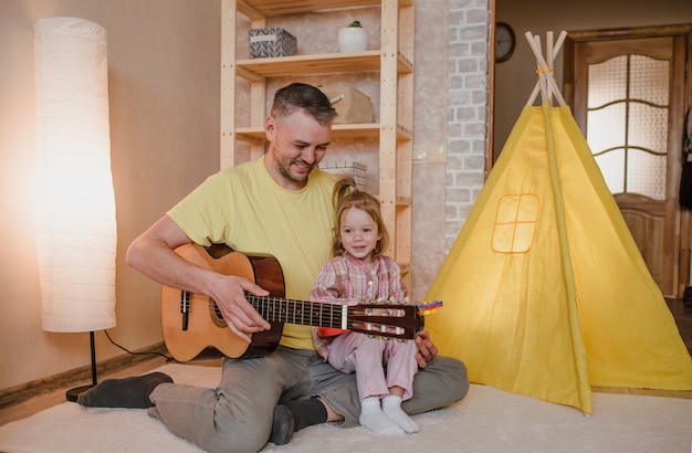 Porträt eines glücklichen Vaters mit einer Gitarre in den Händen und einem kleinen Mädchen, das auf dem Schoß ihres Vaters sitzt. gemeinsame Familienspiele.