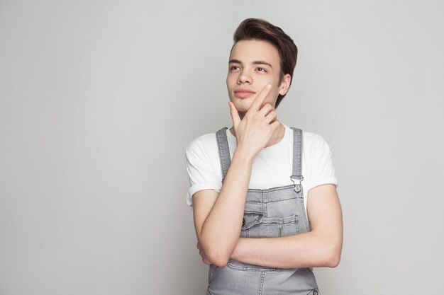 Porträt eines glücklichen, verträumten jungen brünetten Mannes im lässigen Stil mit weißem T-Shirt und Denim-Overalls, der mit Lächeln und Träumen steht und wegschaut. Indoor-Studioaufnahme, auf grauem Hintergrund isoliert.