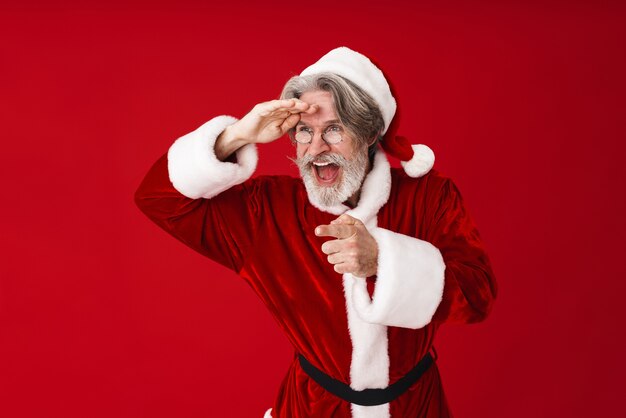 Porträt eines grauhaarigen alten Mannes des Weihnachtsmannes im Kostüm, der mit dem Finger auf das Exemplar einzeln auf Rot schaut und zeigt