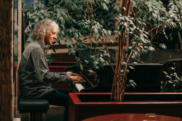 Porträt eines grauhaarigen Senioren mit lockigen langen Haaren sitzt am Klavier und spielt Musik und genießt einen angenehmen Abend im Hotel. Lifestyle-Foto