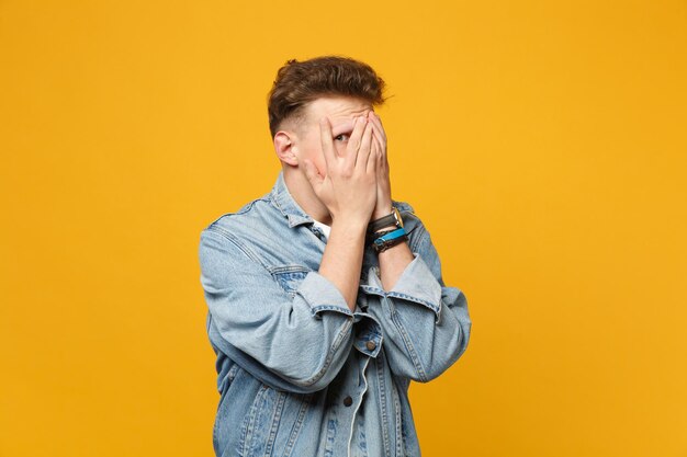 Porträt eines guckenden jungen Mannes in Denim-Freizeitkleidung, der das Gesicht mit den Händen bedeckt, isoliert auf gelb-orangefarbenem Wandhintergrund im Studio. Menschen aufrichtige Emotionen, Lifestyle-Konzept. Mock-up-Kopienbereich.