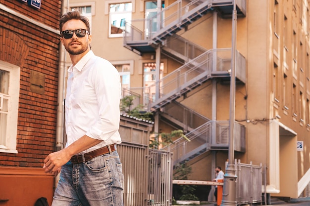 Porträt eines gut aussehenden, lächelnden, stilvollen Hipster-Lambersexual-Modells Moderner Mann in weißem Hemd gekleidet Modemann, der im Straßenhintergrund in der Nähe von Wolkenkratzern mit Sonnenbrille posiert