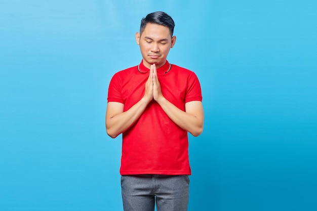 Porträt eines gutaussehenden asiatischen jungen Mannes mit rotem Hemd, der mit den Händen zusammen betet und um Vergebung bittet, isoliert auf blauem Hintergrund