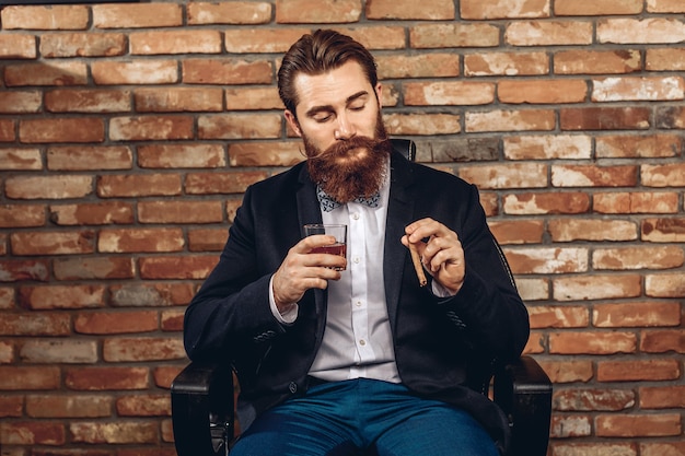 Porträt eines gutaussehenden attraktiven Mannes mit einem Schnurrbart und einem Bart, der auf einem Stuhl sitzt und ein Glas Whisky und eine Zigarre in der Hand hält und gegen eine Mauer posiert. Männliches Sharm-Konzept