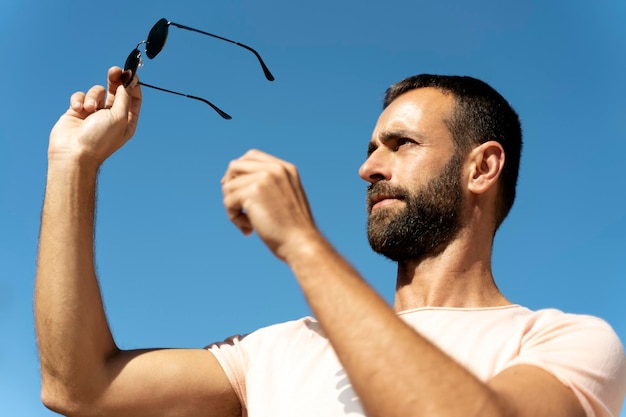 Porträt eines gutaussehenden bärtigen Mannes, der eine stilvolle Sonnenbrille gegen den blauen Himmel hält