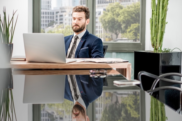 Porträt eines gutaussehenden Bankiers, der mit Laptop im luxuriösen Bürointerieur mit schöner Aussicht auf die Wolkenkratzer arbeitet