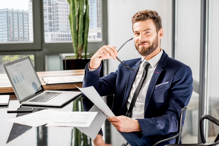 Foto porträt eines gutaussehenden bankiers, der mit laptop im luxuriösen bürointerieur mit schöner aussicht auf die wolkenkratzer arbeitet