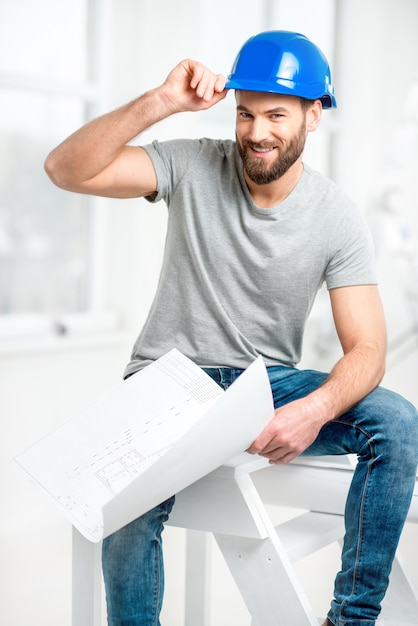 Porträt eines gutaussehenden Baumeisters, Vorarbeiters oder Mechanikers im Helm, der mit Zeichnungen auf Leiter im weißen Innenraum sitzt