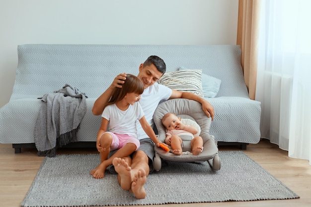 Porträt eines gutaussehenden dunkelhaarigen kaukasischen Mannes, der mit seinen Kindern auf dem Boden sitzt und Glück ausdrückt, Vater kümmert sich und spielt mit seinen Kindern, Vaterschaft und Kindheit.