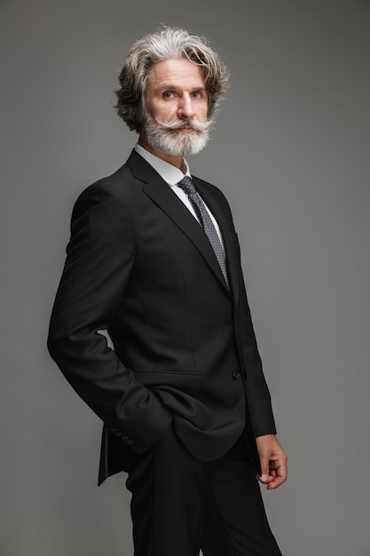 Porträt eines gutaussehenden erwachsenen Geschäftsmannes, der einen formellen schwarzen Anzug trägt, der mit der Hand in der Tasche nach vorne schaut, isoliert über grauer Wand