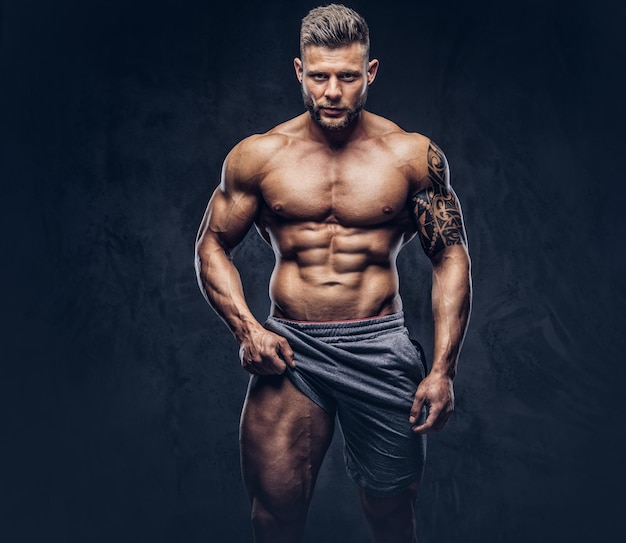 Porträt eines gutaussehenden, hemdlosen, tätowierten Bodybuilders mit stylischem Haarschnitt und Bart, zeigt Muskeln seiner Beine, schaut mit selbstbewusstem Blick in die Kamera und steht in einem Studio. Getrennt auf einem dunklen