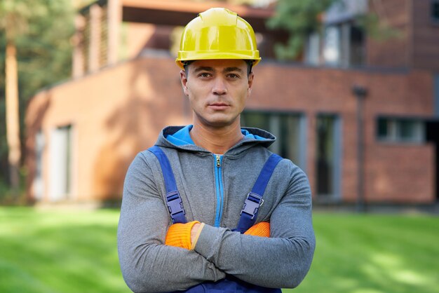 Porträt eines gutaussehenden jungen männlichen Ingenieurs mit Schutzhelm, der in die Kamera schaut und im Freien posiert, während er am Hüttenbau arbeitet. Gebäude, Menschen, umweltfreundliches Baukonzept