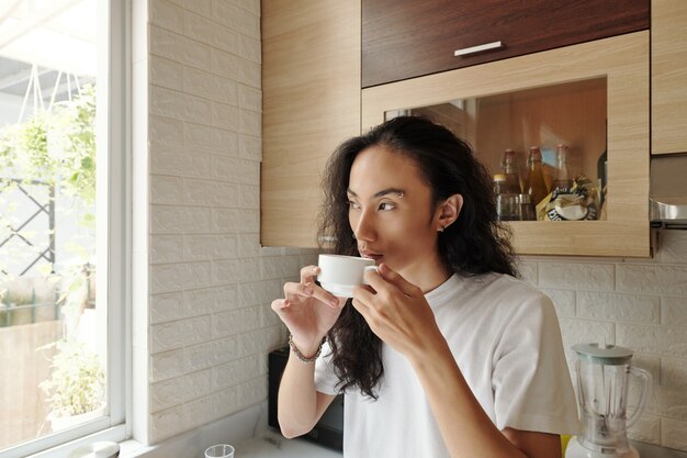 Porträt eines gutaussehenden jungen Mannes, der eine Tasse Morgenkaffee trinkt und durch das Fenster schaut