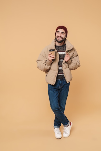 Porträt eines gutaussehenden jungen Mannes in Winterjacke und Hut, der lächelt, während er die Kaffeetasse isoliert auf Beige hält