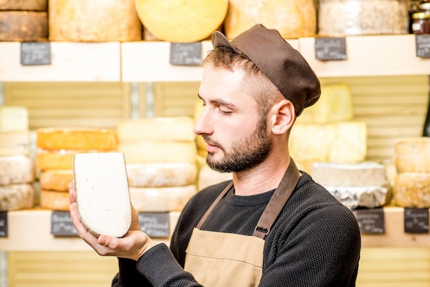 Porträt eines gutaussehenden Käseverkäufers in Uniform, der ein Stück gewürzten Käse vor der Ladenvitrine mit verschiedenen Käsesorten hält