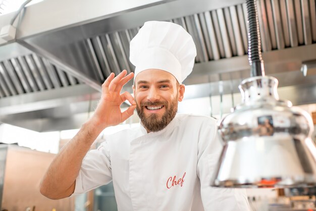 Porträt eines gutaussehenden Kochkochs in Uniform, der ein köstliches Schild in der Restaurantküche zeigt