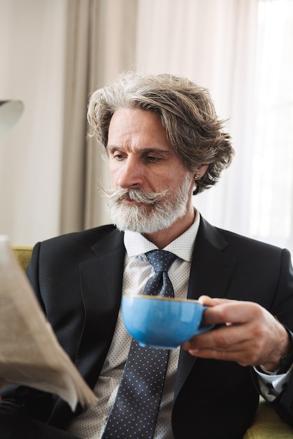 Porträt eines gutaussehenden, konzentrierten, grauhaarigen geschäftsmannes, der zu hause auf einem stuhl sitzt und zeitung trinkt kaffee liest.