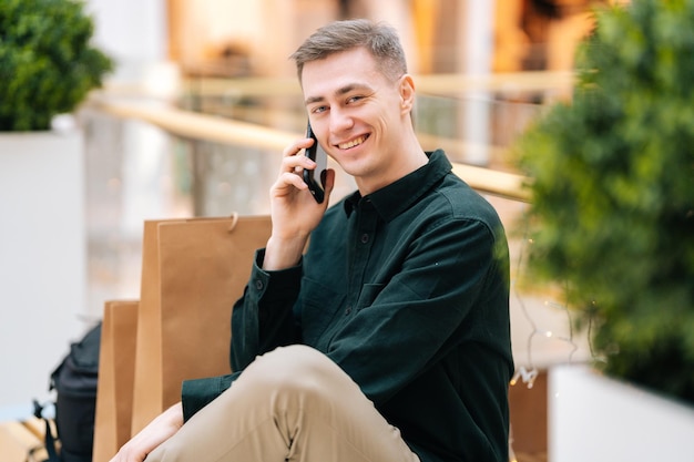 Porträt eines gutaussehenden lächelnden jungen Mannes, der am Handy sitzt und auf einer Bank im Einkaufszentrum sitzt und in die Kamera blickt, selektiver Fokus, unscharfer Hintergrund