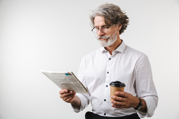 Porträt eines gutaussehenden lächelnden reifen geschäftsmannes in formeller kleidung, der isoliert über weißer wand steht und kaffee zum mitnehmen trinkt, während er eine zeitung liest