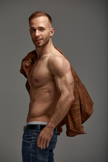 Porträt eines gutaussehenden männlichen Models, das in einer braunen Lederjacke und Jeans ohne Hemd posiert Grauer Hintergrund Modische Herbstkleidung Herren-Jugendmode