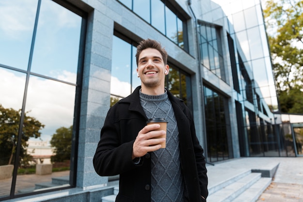 Porträt eines gutaussehenden Mannes der 30er Jahre, der Ohrstöpsel trägt und eine Pappbecher Kaffee zum Mitnehmen hält, während er vor einem Glasgebäude schlendert