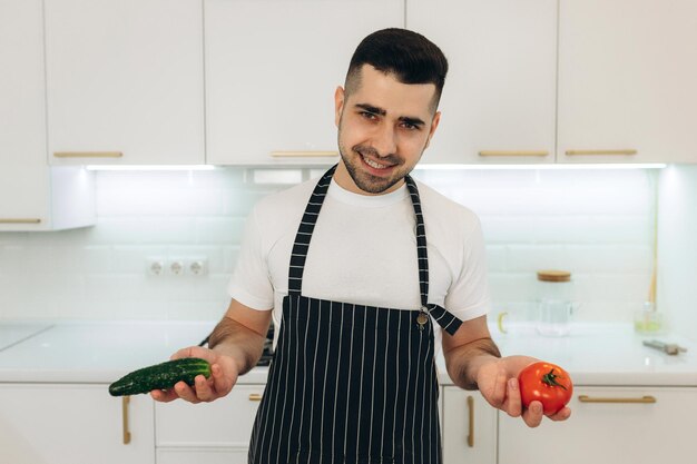 Porträt eines gutaussehenden Mannes in der Küche Der Typ trägt eine schwarze Schürze In seiner Hand hält er eine rote Tomate und eine grüne Gurke