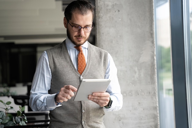 Porträt eines gutaussehenden Mannes mittleren Alters, der ein digitales Tablet in der Hand hält und glücklich in die Kamera lächelt, die am Fenster in einem modernen Büro steht