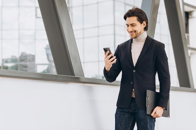 Porträt eines gutaussehenden modernen männlichen Geschäftsmannes im Anzug, der einen Ordner mit Dokumenten hält und vor dem Hintergrund städtischer Gebäude und Büros telefoniert