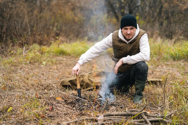 Porträt eines gutaussehenden Reisenden, der warme Kleidung trägt und in der Nähe eines brennenden Lagerfeuers sitzt und eine kleine Schaufel hält, die in die Kamera schaut