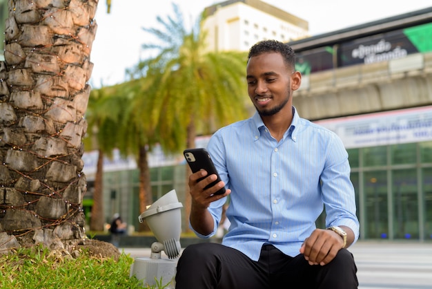 Porträt eines gutaussehenden schwarzafrikanischen Geschäftsmannes im Freien in der Stadt während des Sommers, der im Sommer sitzt und Handy benutzt, während er horizontal lächelt