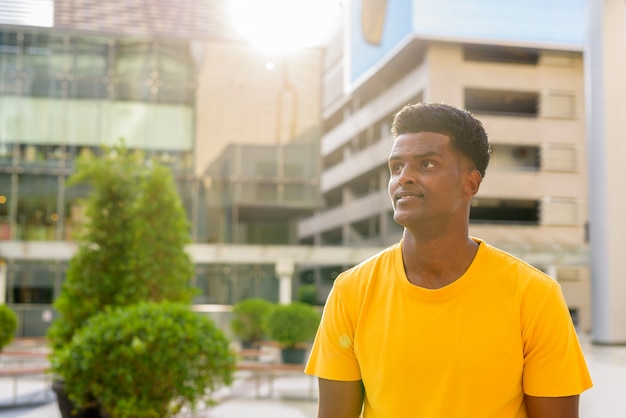 Porträt eines gutaussehenden schwarzen afrikanischen Mannes mit gelbem T-Shirt im Freien in der Stadt während des Sommers mit Hintergrundbeleuchtung mit Lens Flare beim Denken