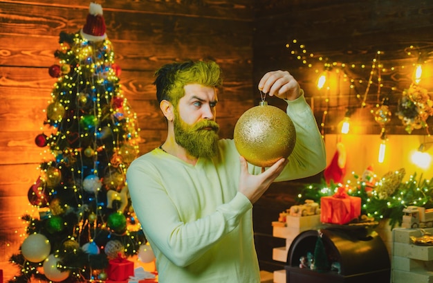 Porträt eines gutaussehenden Weihnachtsmanns drinnen mit Weihnachtskugel, Goldkugel, bärtiger Mann, der in der Nähe Spaß hat