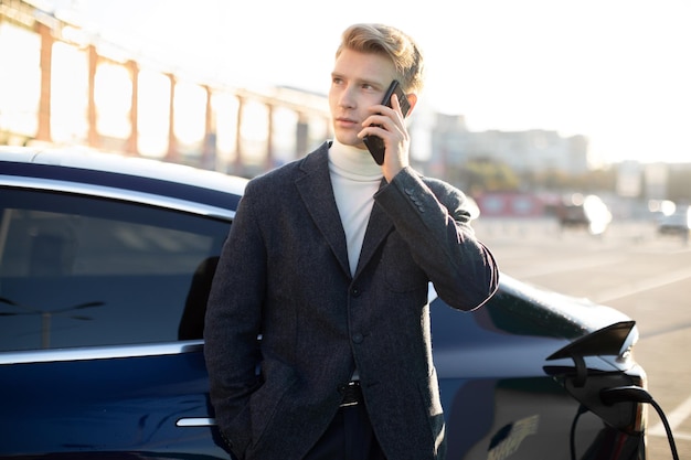 Porträt eines gutaussehenden, zielstrebigen jungen Geschäftsmannes, der wichtige Anrufe und Gespräche führt, während sein Auto an der Ladestation für Elektrofahrzeuge aufgeladen wird
