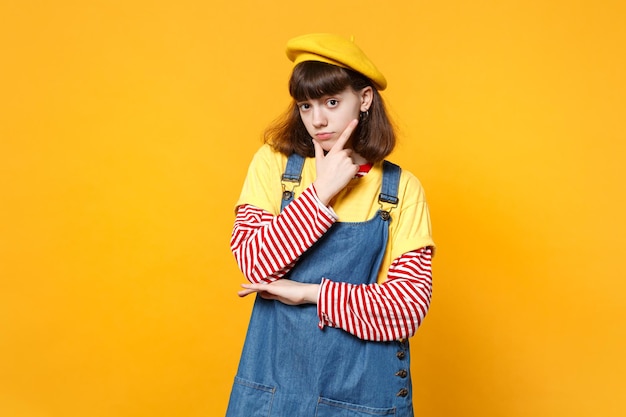 Porträt eines hellen Mädchen-Teenagers in französischer Baskenmütze, Denim-Sommerkleid, Handstütze auf Kinn gelegt, isoliert auf gelbem Wandhintergrund im Studio. Menschen aufrichtige Emotionen, Lifestyle-Konzept. Mock-up-Kopienbereich.