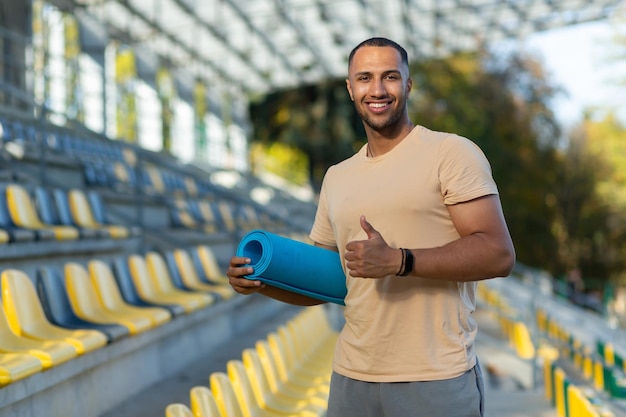 Porträt eines hispanischen männlichen Fitnesstrainers im Sportstadion mit lächelnder und lächelnder Yogamatte