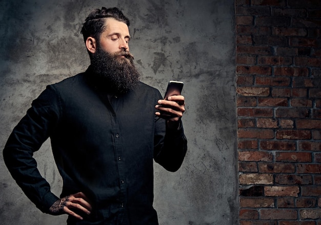 Porträt eines hübschen bärtigen Hipsters in einem schwarzen Hemd, der ein Smartphone benutzt und in einem Studio steht. Getrennt auf einem dunklen Hintergrund.