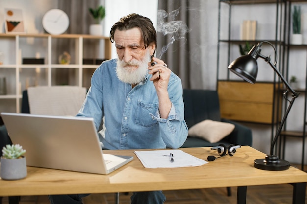 Porträt eines hübschen, eleganten, reifen Geschäftsmanns mit Zigarre, der mit Laptop am Schreibtisch sitzt