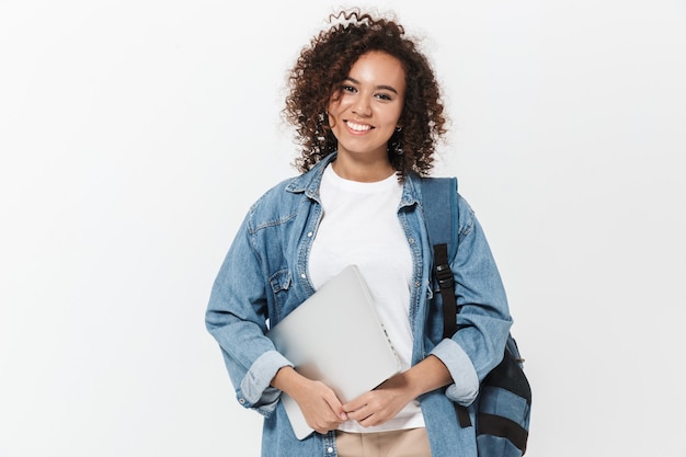 Porträt eines hübschen fröhlichen, lässigen afrikanischen Mädchens, das einen Rucksack trägt, der isoliert über der weißen Wand steht und einen Laptop hält?