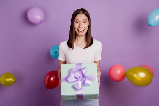 Porträt eines hübschen, funky fröhlichen Mädchens, das Luftballons in den Händen hält, die Geschenkbox einzeln auf lebendigem violettem Farbhintergrund hält