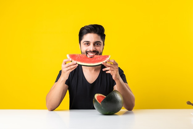 Porträt eines hübschen indischen jungen Mannes, der lächelt und frische Wassermelone oder Tarbooj isst, während er am Tisch sitzt oder über Holzboden isoliert