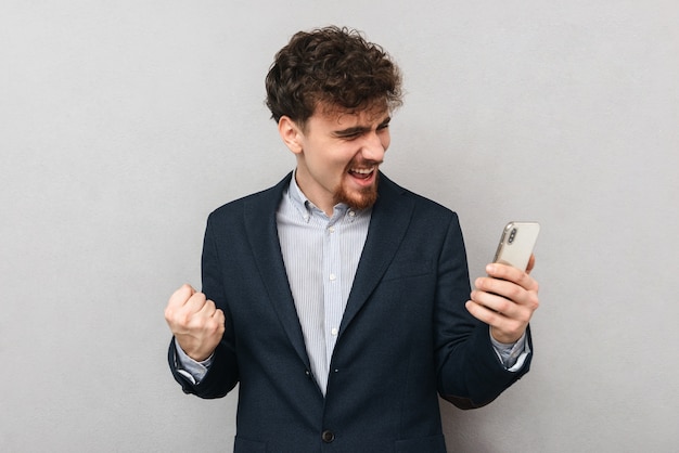 Porträt eines hübschen jungen Geschäftsmannes lokalisiert über graue Wand machen Gewinnergeste unter Verwendung des Mobiltelefons.