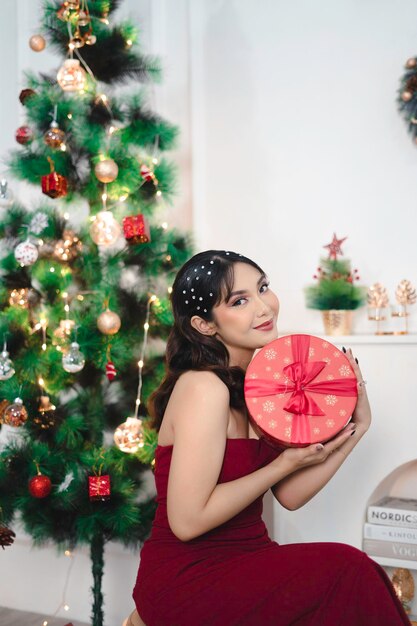 Porträt eines hübschen jungen Mädchens, das sich gemütlich hinsetzt und ein Weihnachtsgeschenk hält, das ein rotes Kleid im geschmückten Weihnachtswohnzimmer im Innenbereich trägt