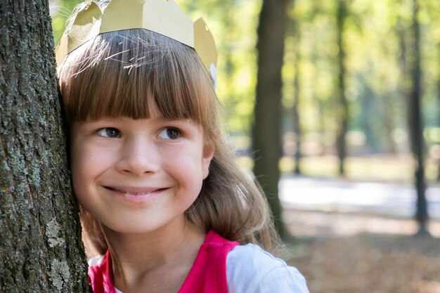 Porträt eines hübschen lächelnden Kindermädchens in einer Papierkrone auf ihrem Kopf, das an einen Baum im Sommerpark lehnt.