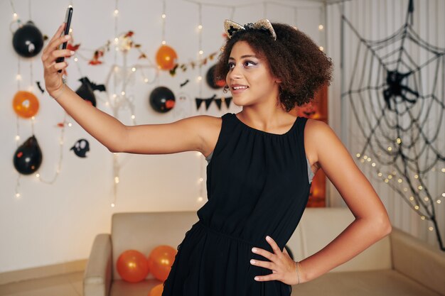 Porträt eines hübschen lächelnden Teenager-Mädchens mit Katzenohren, das Selfie auf der Halloween-Party macht